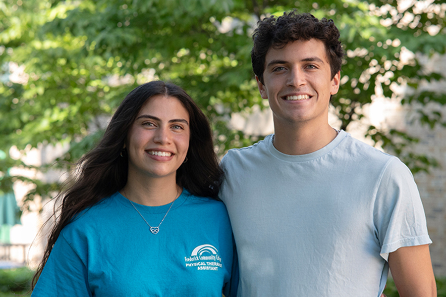 A young man and woman are smiling and posing for a photo together outdoors. The woman on the left has long, dark, wavy hair and is wearing a blue t-shirt with a Frederick Community College Physical Therapist Assistant logo on the chest. The man on the right has short, curly, dark hair and is wearing a light blue t-shirt. They are standing in front of a background of lush green trees and foliage.