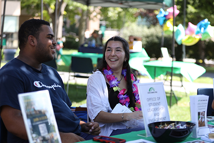 Two students smiling and talking at an outdoor "Student Life office " information table.